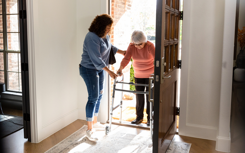woman helping female patient with walker through front door 