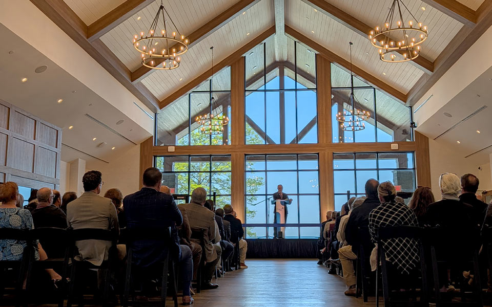 Audience seated in a large timber-framed event hall listening to a speaker at a podium